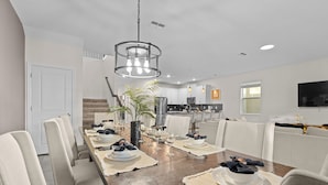 Comedor con una larga mesa de madera dispuesta para seis personas, rodeada de sillas tapizadas. Un candelabro moderno cuelga encima, y se ve una cocina al fondo.