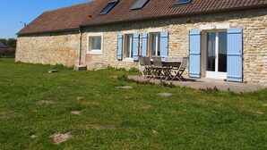 A stone building with a tiled roof, a patio with a table and chairs, and a grassy yard.