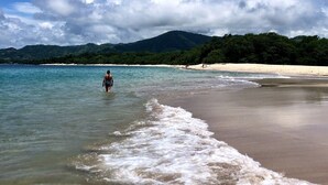 Una playa con agua cristalina, una persona caminando y una montaña al fondo.