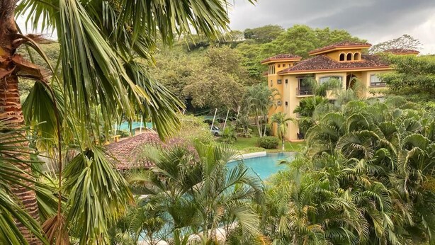 Una piscina rodeada de exuberante vegetación y un edificio amarillo con tejado de baldosas.