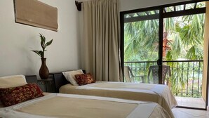 Habitación de hotel con dos camas, una planta en maceta, una ventana con vistas a palmeras y un balcón con sillas.