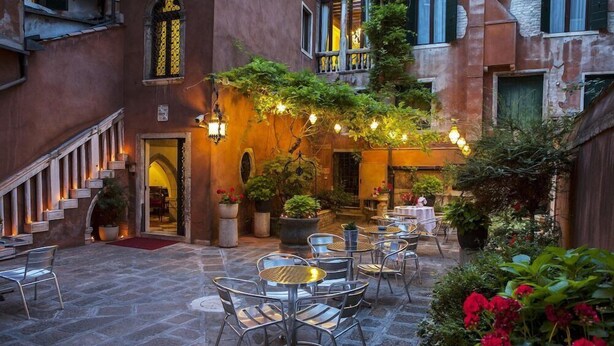 An outdoor café area with stone flooring, metal chairs, and tables, surrounded by potted plants and a staircase leading to an entrance. - San Moise Hotel (Venice)