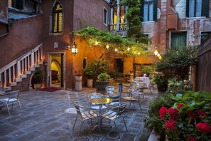 An outdoor café area with stone flooring, metal chairs, and tables, surrounded by potted plants and a staircase leading to an entrance. - San Moise Hotel (Venice)