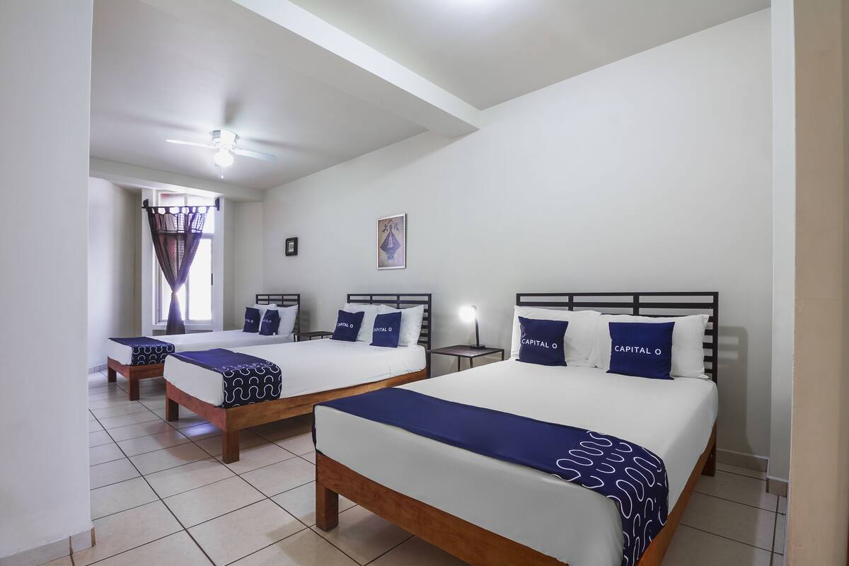 A hotel room with two beds, a ceiling fan, a window with curtains, and a small table with a lamp.