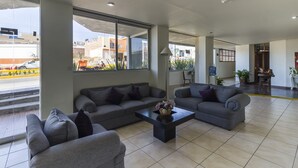 A modern living area with grey sofas and a coffee table, large windows offering a view of the street and buildings, and a clean, tiled floor. - Capital O Plaza Galindo (San Juan de los Lagos)
