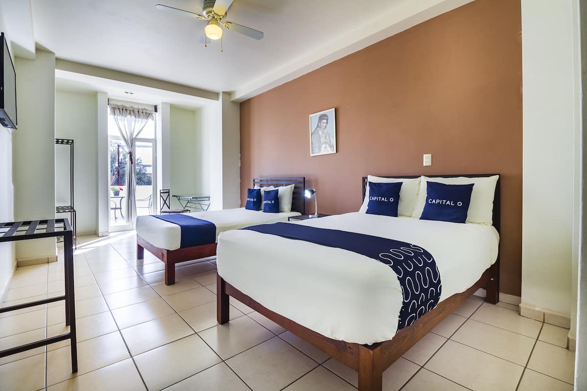 A hotel room with two beds, a ceiling fan, a large window with curtains, a framed picture on the wall, and a small table with a chair.