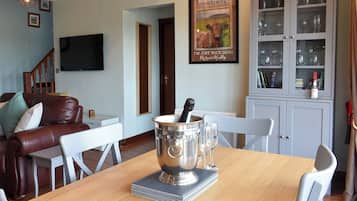 A dining area with a wooden table, white chairs, a silver cake stand, and a bottle of champagne. There is a framed picture on the wall and a cabinet with glass doors containing glasses.