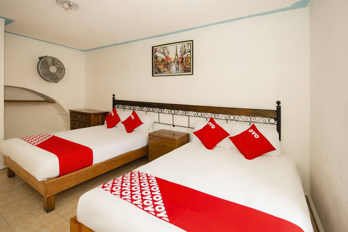 A hotel room with two beds, each with a red and white bedspread and matching pillows. There is a small wooden nightstand between the beds, and a wall-mounted fan above the left bed. A framed picture of a city street hangs on the wall above the beds.