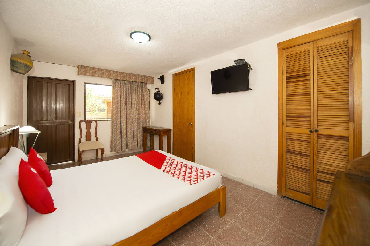 A bedroom with a bed, a wooden wardrobe, a TV, and a window with curtains.
