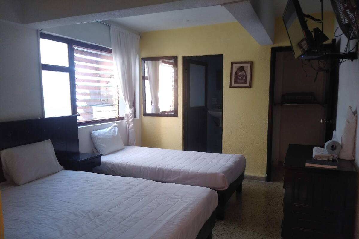 A room with two beds, a window with curtains, a mirror, a framed picture on the wall, and a desk with a lamp.