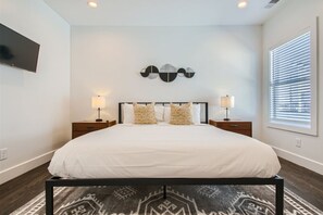 Une chambre à coucher avec un grand lit, deux tables de chevet avec des lampes, un téléviseur fixé au mur et une fenêtre avec des stores.