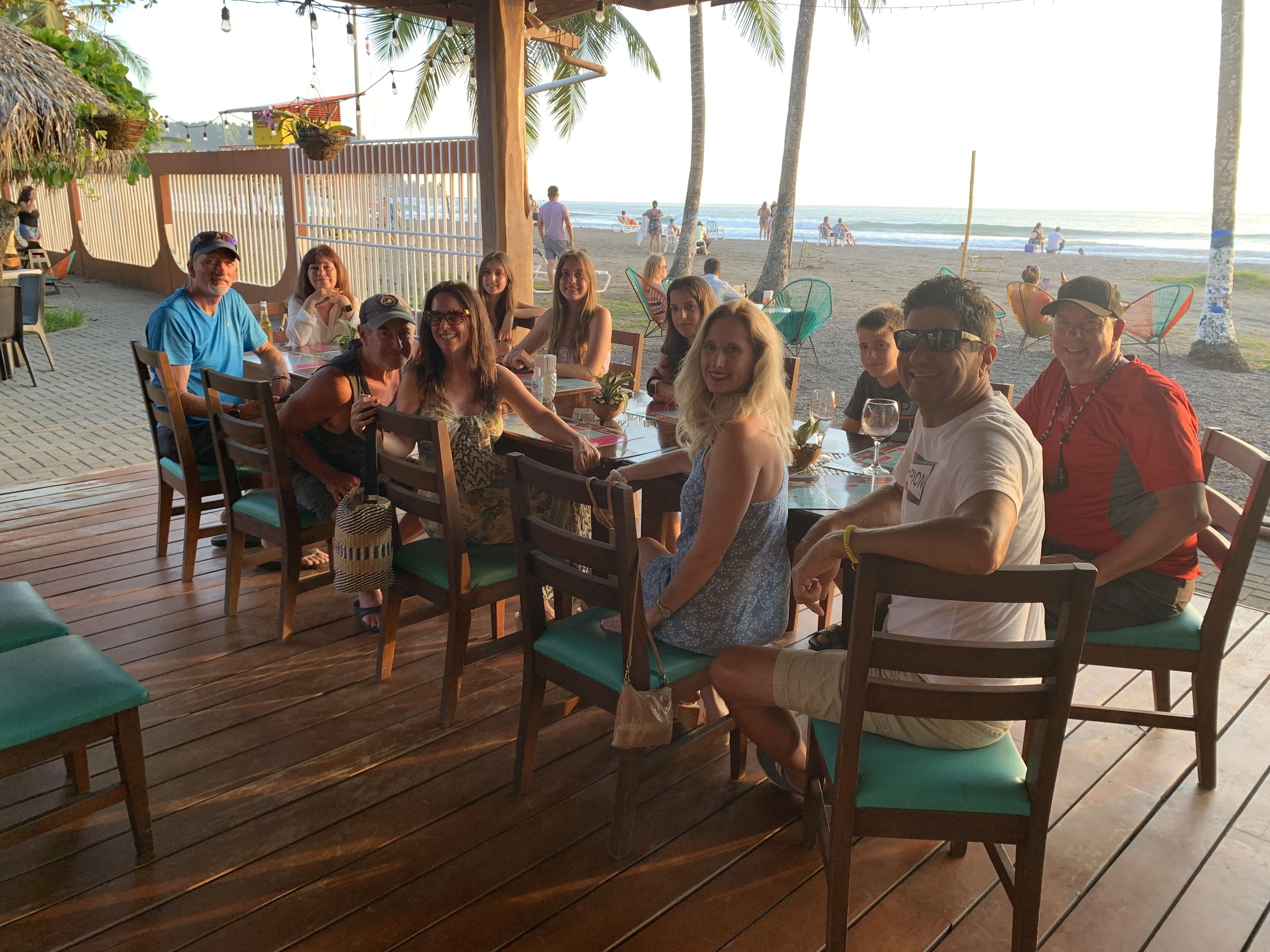 Een groep mensen zit aan tafels op een houten terras, met uitzicht op het strand en de oceaan.