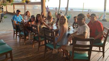 Een groep mensen zit aan tafels op een houten terras, met uitzicht op het strand en de oceaan.