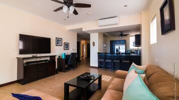A living room with a TV, sofa, coffee table, and ceiling fan. There is a kitchen in the background.