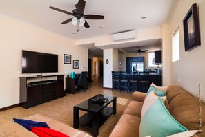 A living room with a TV, sofa, coffee table, and ceiling fan. There is a kitchen in the background. - Ocean front and just steps from the pools and ocean! (Jacó)