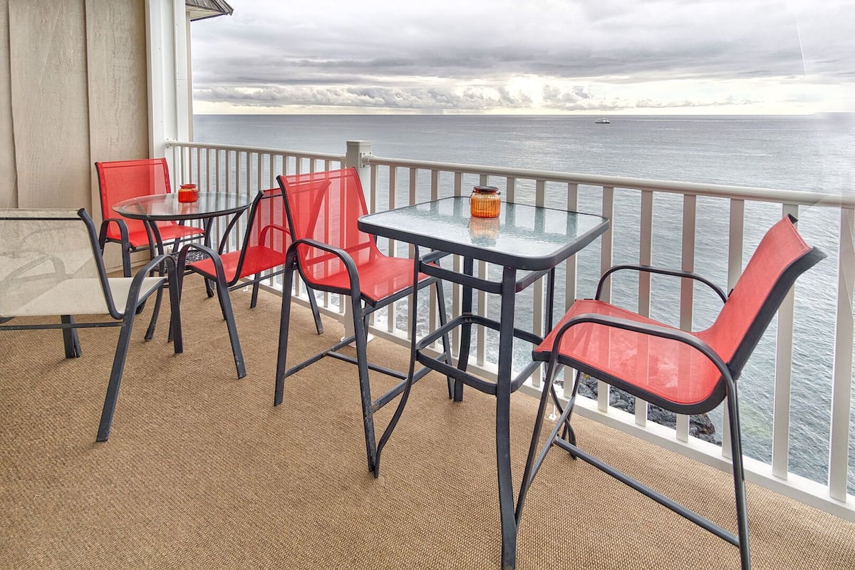 Ein Balkon mit Tisch und Stühlen, mit Blick auf ein Gewässer und einen bewölkten Himmel.