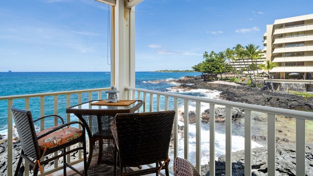 A balcony with wicker chairs and a small table, overlooking a rocky coastline and ocean.