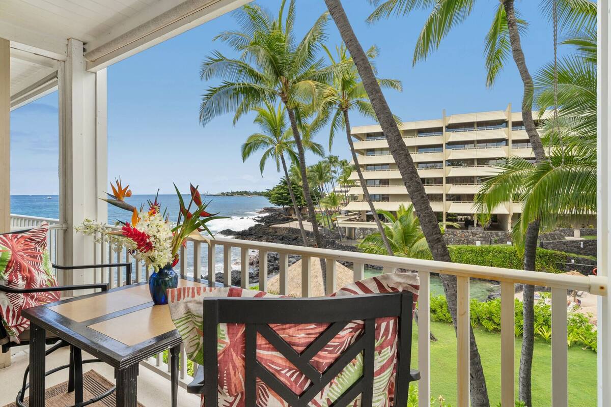 Ein Balkon mit Tisch und Stühlen, Blick auf einen Strand mit Palmen und ein Gebäude im Hintergrund.