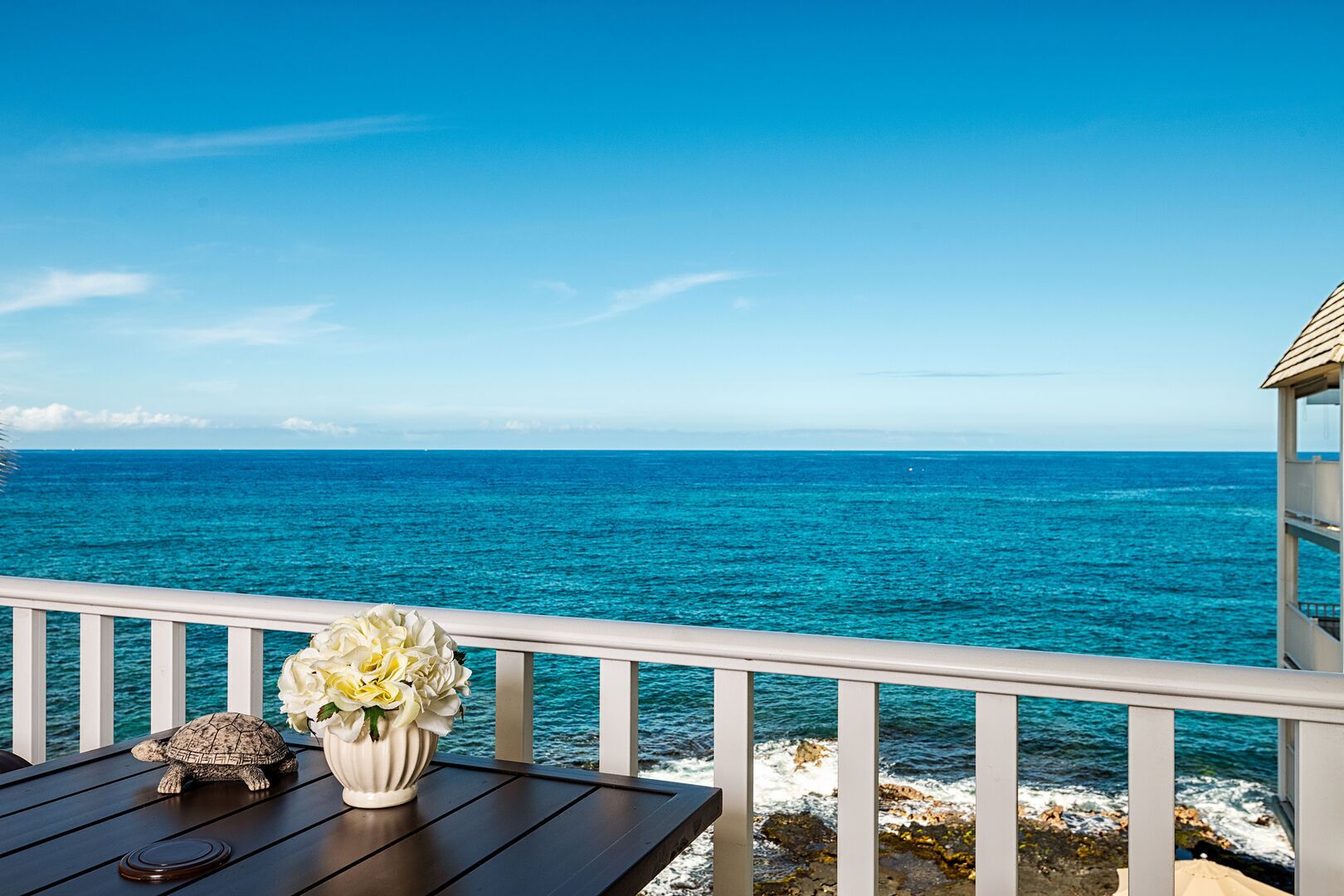 Ein Balkon mit Blick auf das Meer, einem weißen Geländer und einem Tisch mit einer Schildkrötenfigur und einem Blumenstrauß.