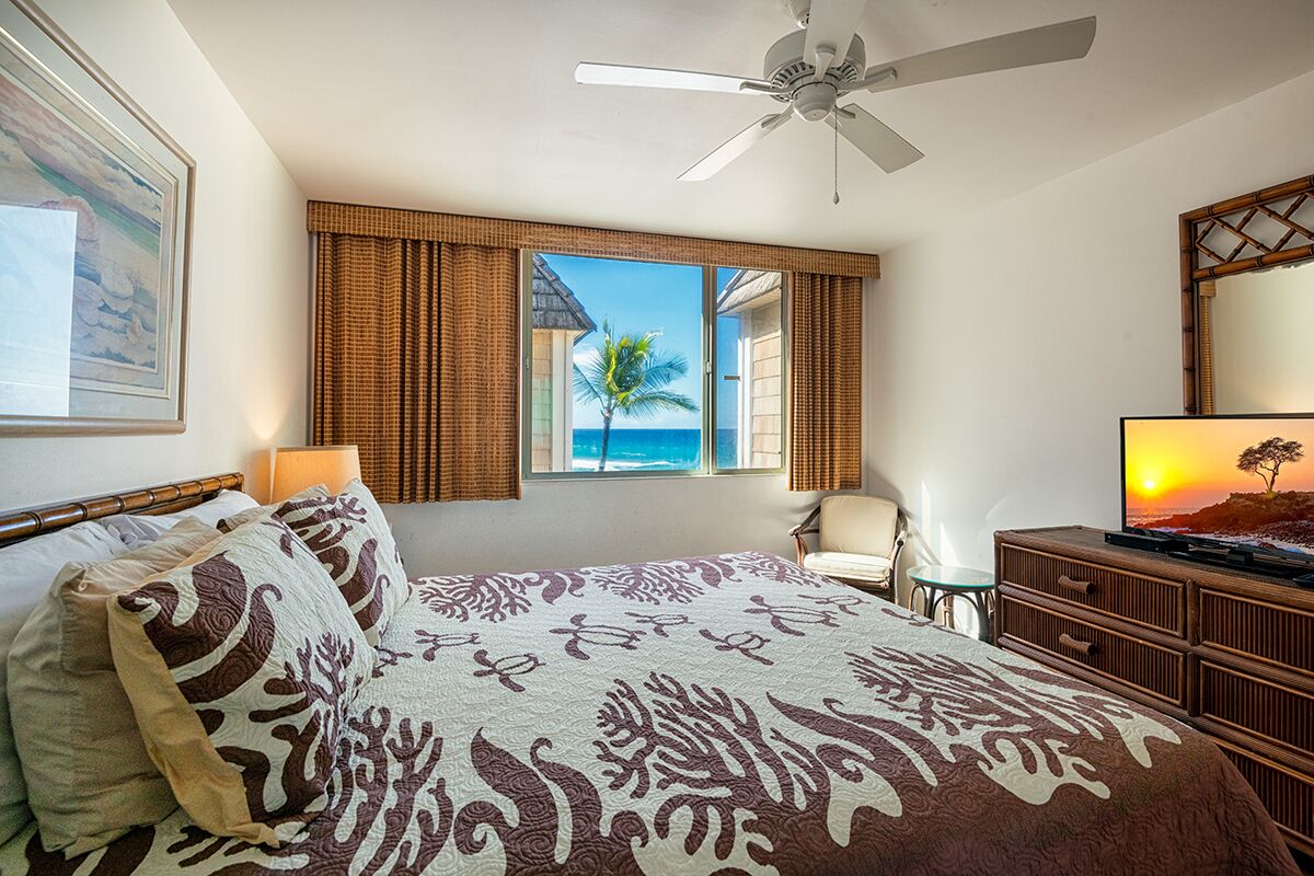 Un dormitorio con cama, ventilador de techo, televisor, ventana con vista al océano y una silla.