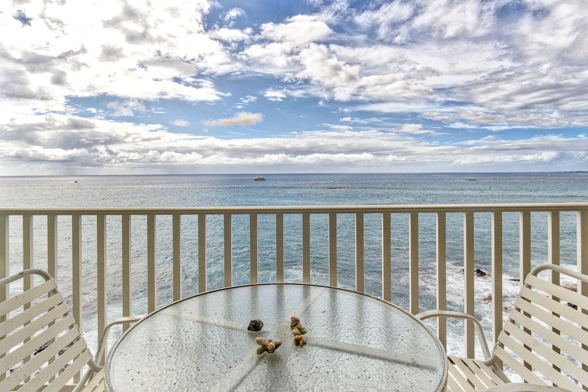 Ein Balkon mit einem Glastisch und weißen Stühlen, der auf ein ruhiges Meer mit einem Schiff in der Ferne und einem Hund am Ufer blickt.
