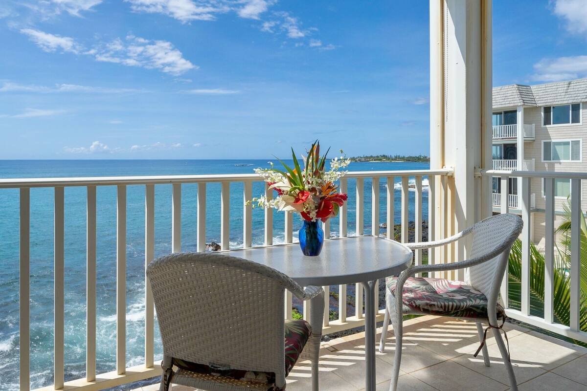 Ein Balkon mit Blick auf das Meer, ein Tisch mit Blumenvase und zwei Korbstühle.