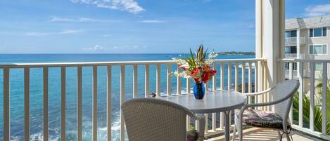 A balcony with a view of the ocean, a table with a vase of flowers, and two wicker chairs.