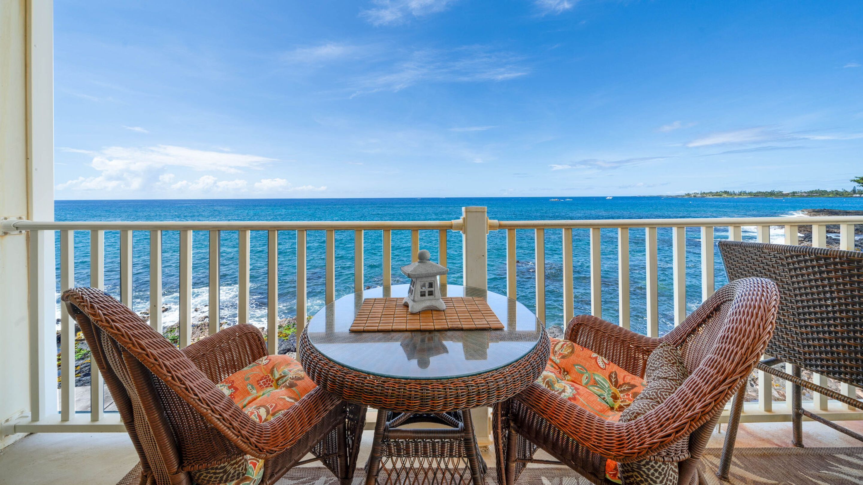 A balcony with wicker furniture and a table, overlooking a coastal view.