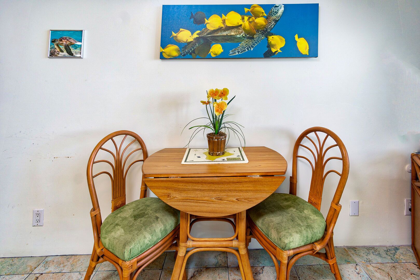 A dining area with a round wooden table, two wicker chairs with green cushions, and a small potted plant on a placemat.