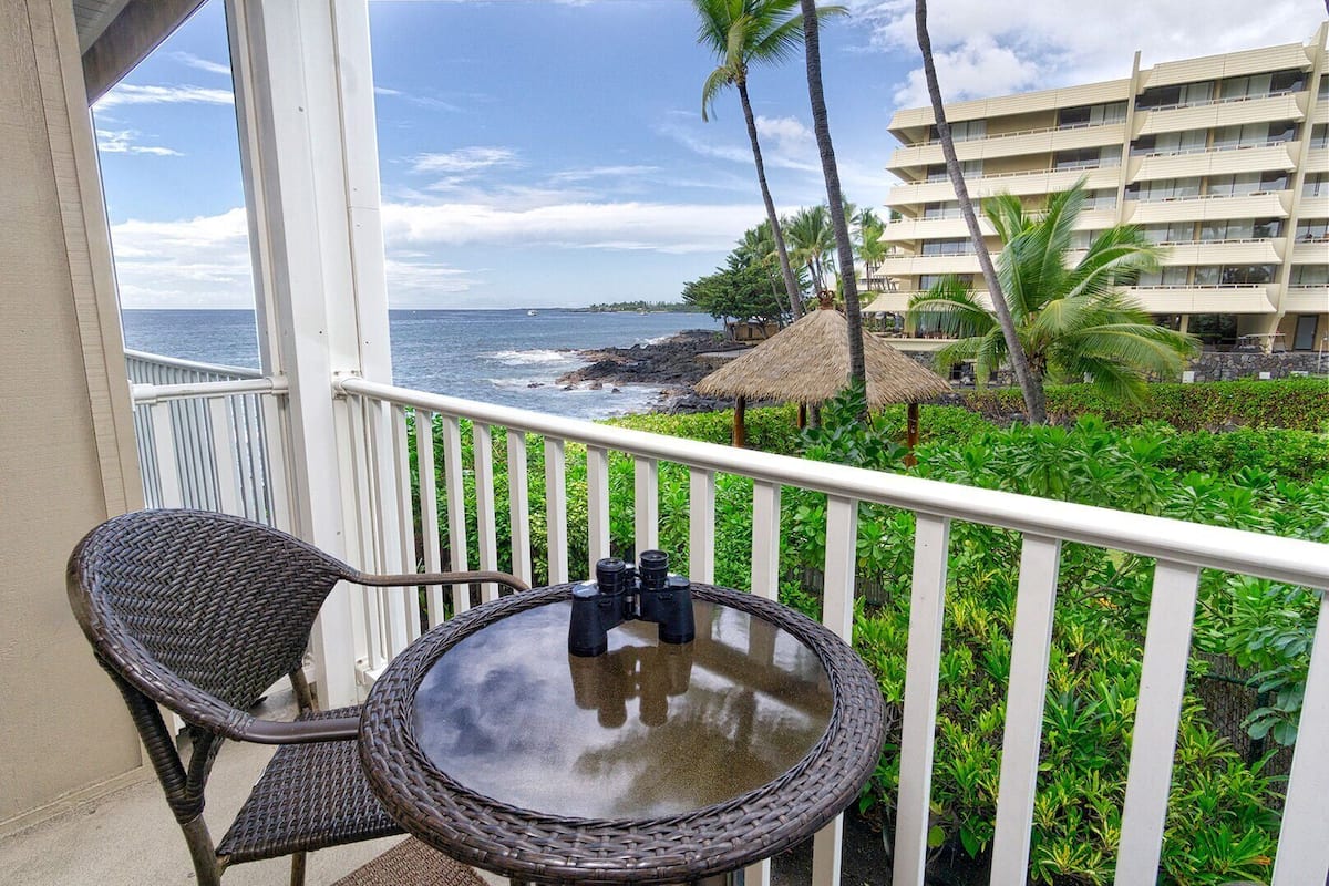 Ein Balkon mit Korbstühlen und einem Tisch, der Blick auf einen Strand mit Palmen und ein Hotelgerichtetes Gebäude freigibt.