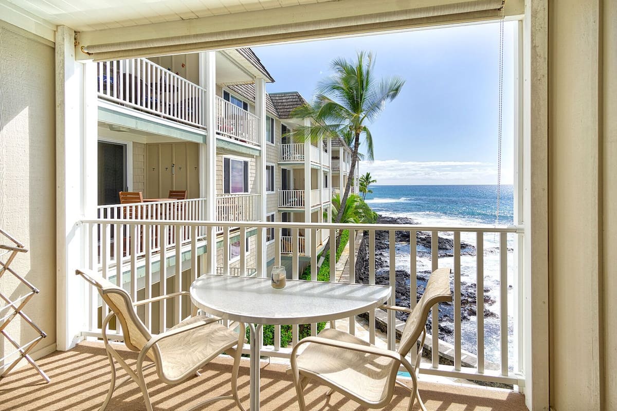 Ein Balkon mit Tisch und Stühlen, der Blick auf einen Strand mit Palmen und den Ozean freigibt.