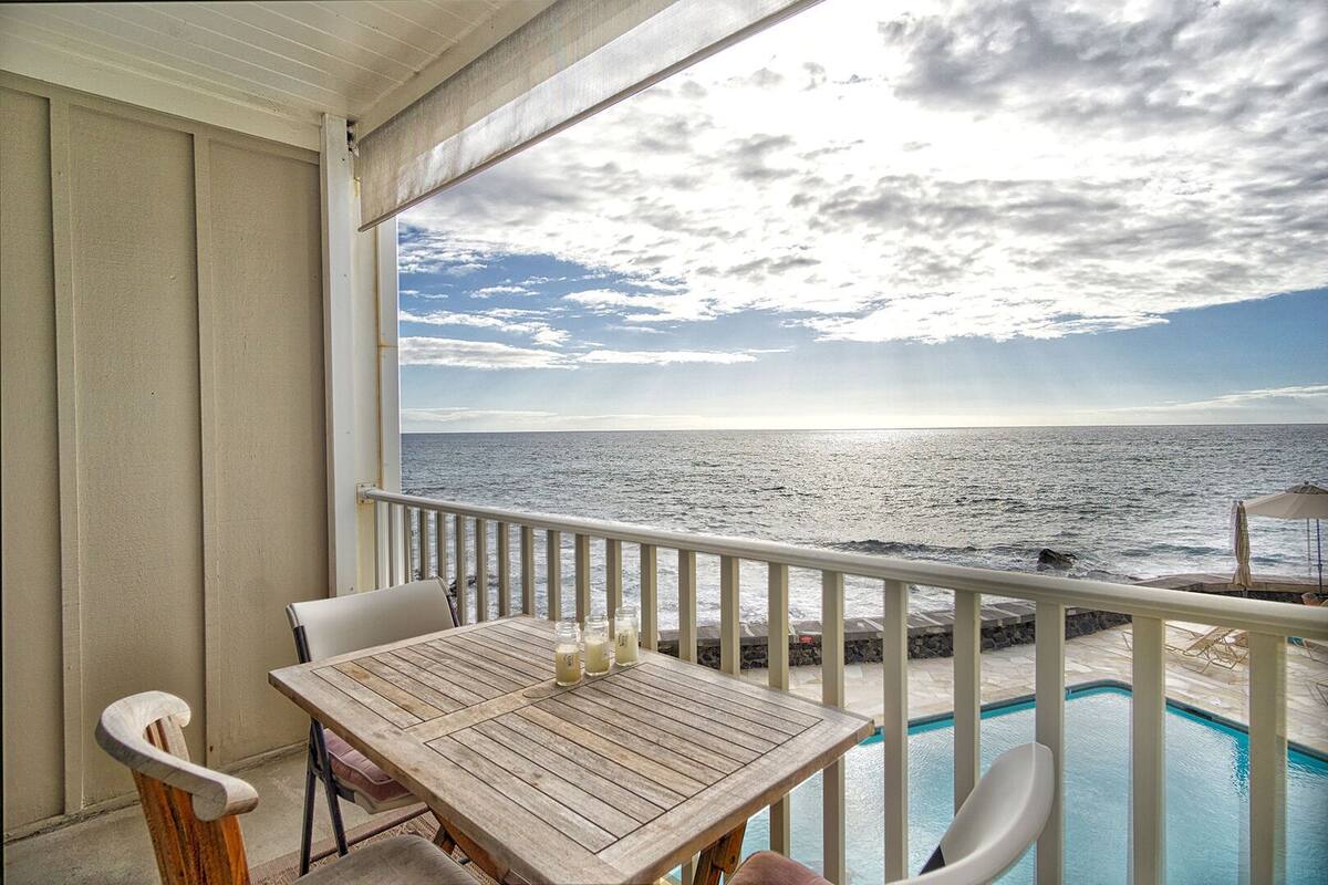 Ein Balkon mit Holztisch und Stühlen mit Blick auf einen Swimmingpool und den Ozean.