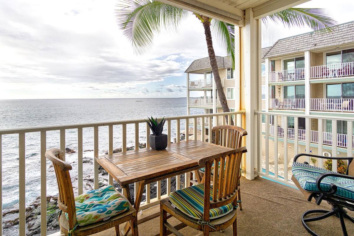 Ein Holztisch mit Stühlen auf einem Balkon mit Blick auf das Meer und ein Gebäude mit Balkonen.