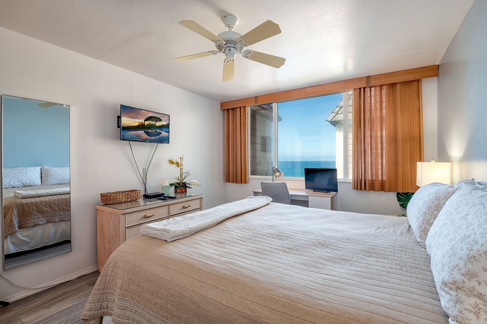 A hotel room with a bed, a desk, a TV, a ceiling fan, and a large window with curtains.