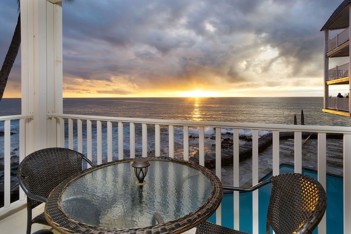 Ein Balkon mit Glastisch und Korbstühlen, mit Blick auf einen Sonnenuntergang über dem Meer.