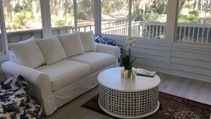 Un sofá blanco con cojines azules, una mesa redonda blanca con velas, y vistas a una terraza de madera y vegetación.