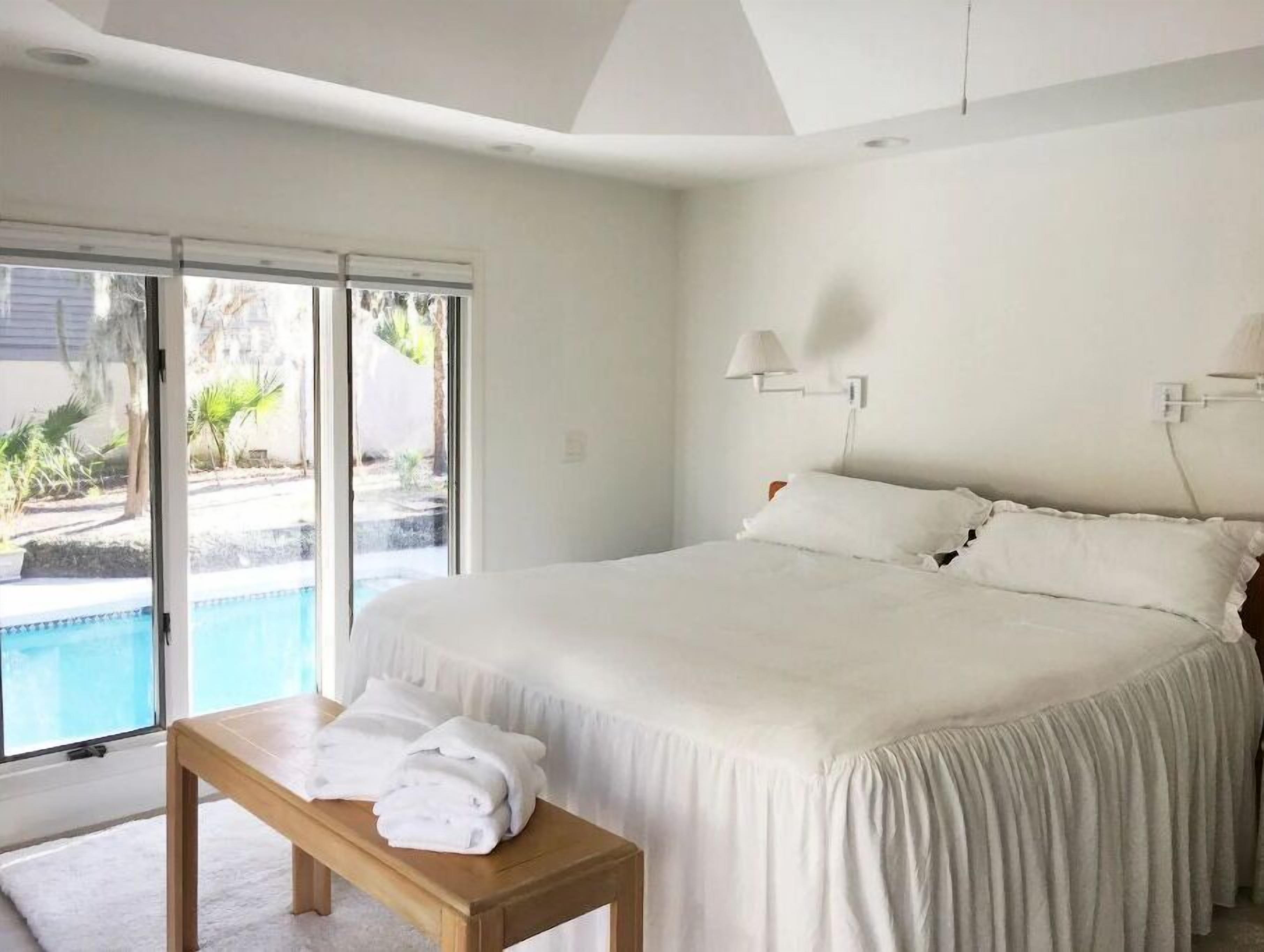 Ein Schlafzimmer mit einem großen Bett, einer Bank mit Handtüchern, einem Fenster mit Blick auf Pool und Grünflächen und einer Deckenleuchte.