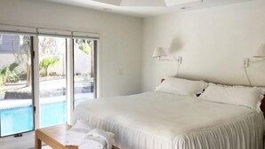Un dormitorio con una cama grande, un banco con toallas, una ventana con vista a una piscina y follaje, y una lámpara en el techo.