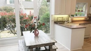Una cocina con un ventanal grande, una mesa de comedor con un florero y armarios blancos.