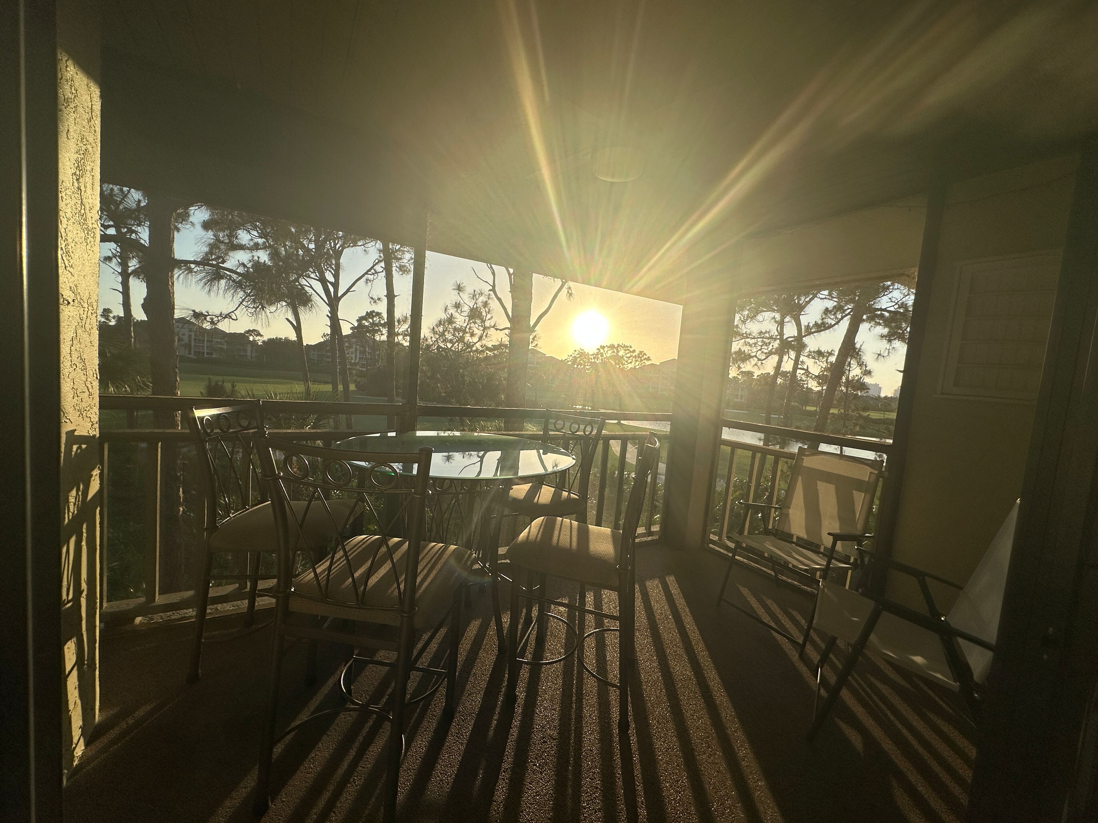 A sunlit outdoor patio with a round table and chairs, overlooking a body of water and trees.