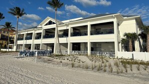 Un hôtel à plusieurs étages, doté de balcons, situé sur une plage de sable fin bordée de palmiers.