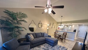 Un salon avec un canapé d’angle, une petite table ronde, un fauteuil bleu, une grande plante en pot, un ventilateur de plafond et, au fond, une cuisine.