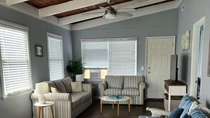 A spacious living room with a high ceiling, wooden beams, and large windows with blinds. There are two sofas, a coffee table, and a television.