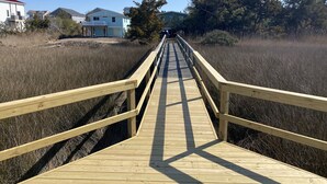 A wooden boardwalk leading to a house with a green roof, surrounded by tall grass and trees.