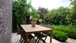 An outdoor patio with a wooden table and chairs, surrounded by lush greenery and a brick wall.