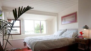 A bedroom with a wooden bed, a nightstand with a lamp, a framed picture on the wall, and a window with a view of greenery outside.