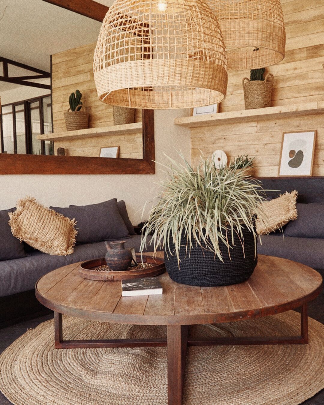 Un salon comprenant une table basse ronde en bois, une petite table d’appoint en osier de couleur sombre, un pouf en osier et une grande plante en pot.