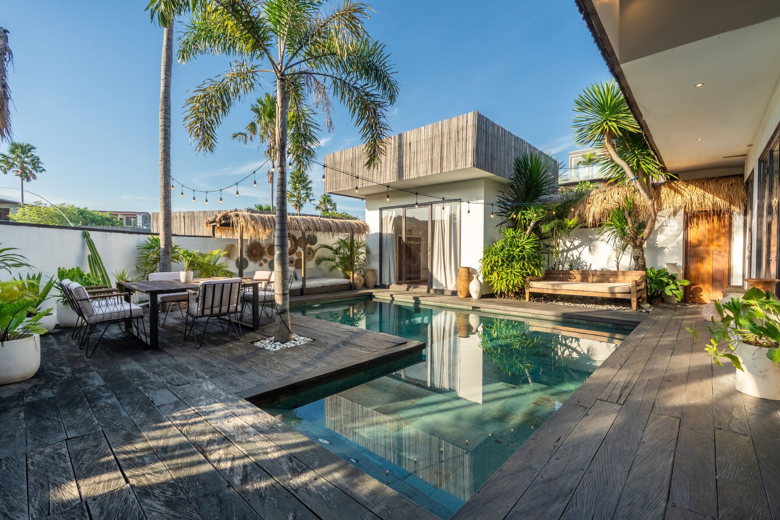 Een modern buitenzwembad met een houten terras, een eethoek en tropische planten.