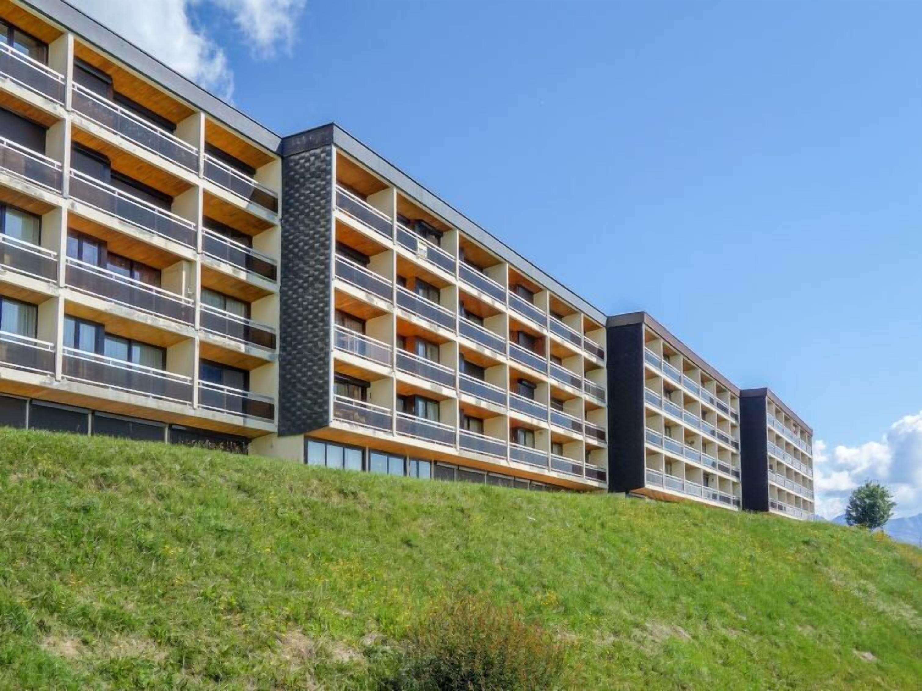 A multi-story hotel with balconies, a modern architectural design, and a grassy hill in the foreground.