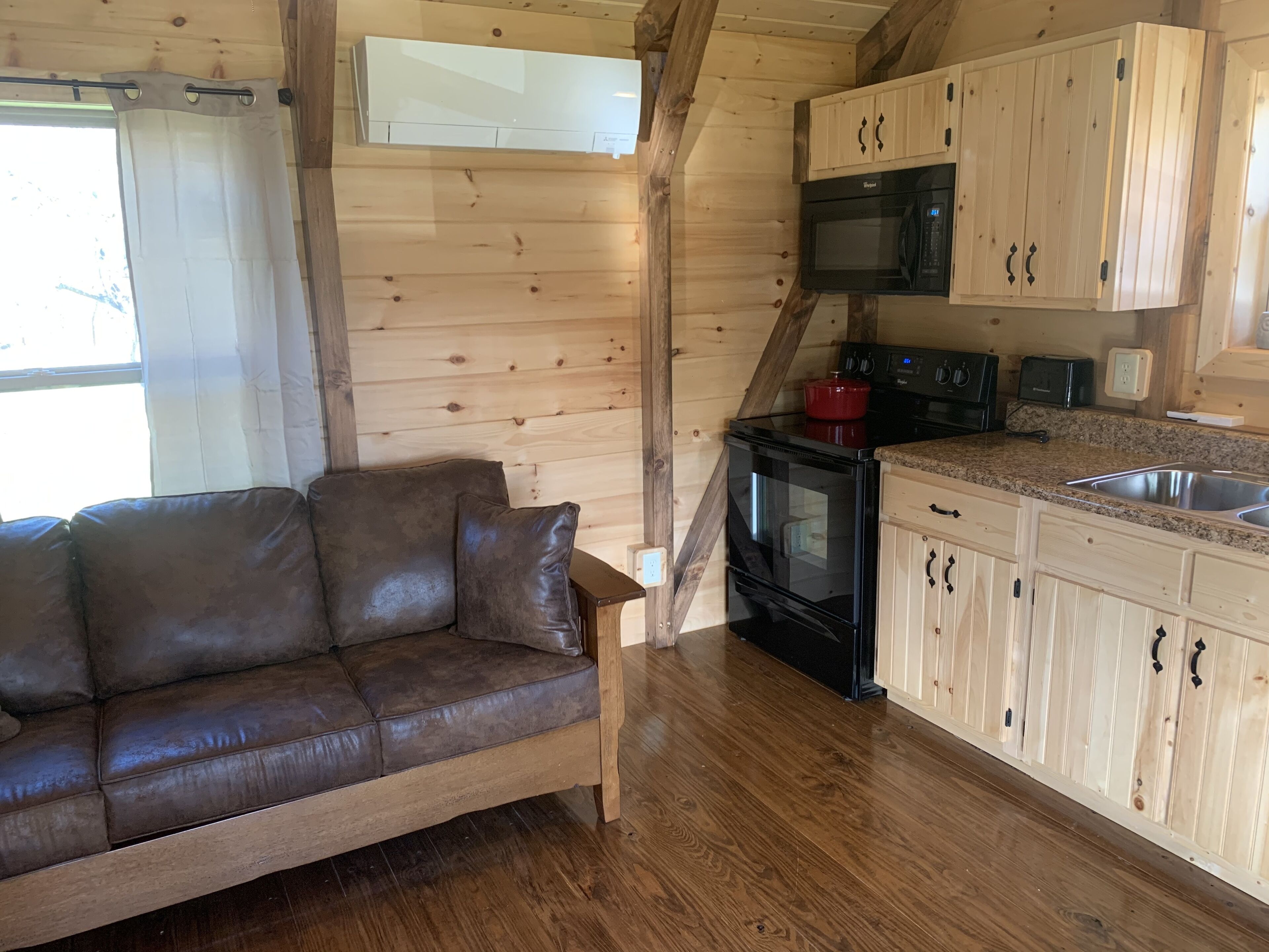 Interior acogedor de una cabaña con piso de madera, un sofá de cuero marrón, una zona de cocina con gabinetes de madera, un microondas, una estufa y una olla roja.